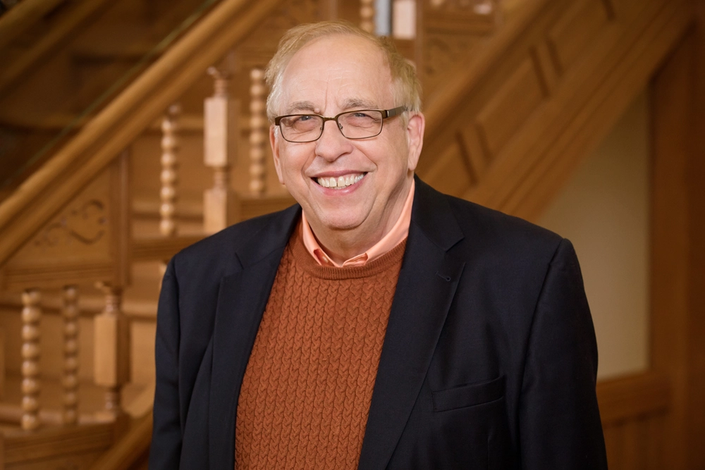 A person in a blazer with a sweater underneath standing in front of a wooden staircase